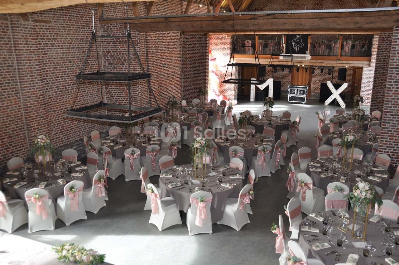 Salle de réception décorée pour un mariage, avec des tables rondes dressées, chaises ornées de nœuds roses et fleurs.