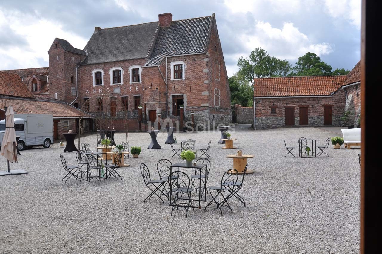 Cour pavée avec tables et chaises en métal, entourée de bâtiments en briques anciennes sous un ciel nuageux.