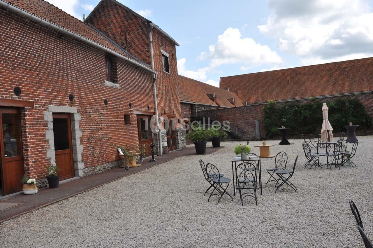 Cour pavée avec mobilier de jardin en fer forgé, entourée de bâtiments en briques rouges sous un ciel partiellement nuageux.