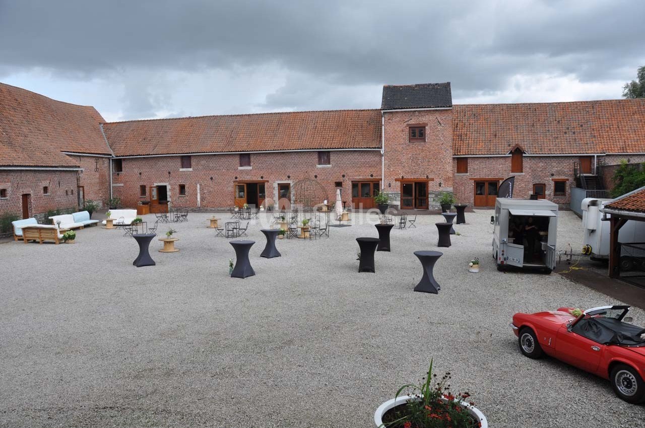 Cour intérieure d'une ferme rénovée avec tables hautes, mobilier de réception et une voiture ancienne rouge.