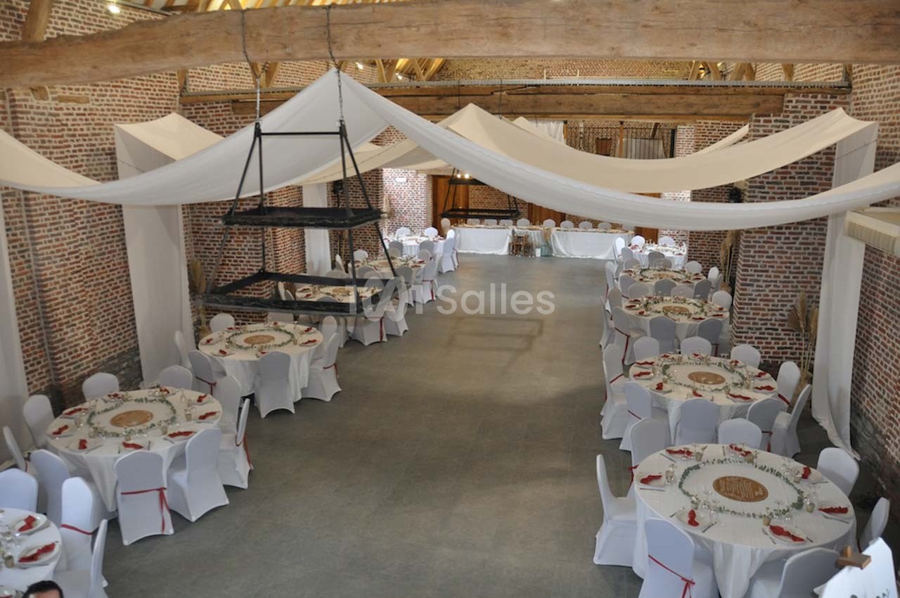 Salle de réception avec murs en briques, poutres apparentes, tables rondes dressées et décorations en tissu suspendues.