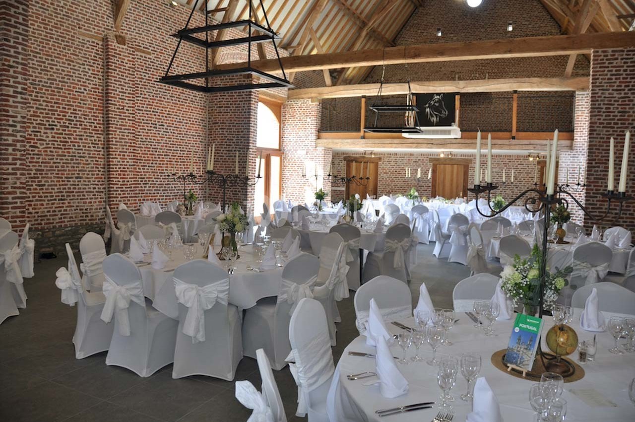 Salle de réception avec murs en briques, poutres apparentes et tables rondes dressées pour un événement.
