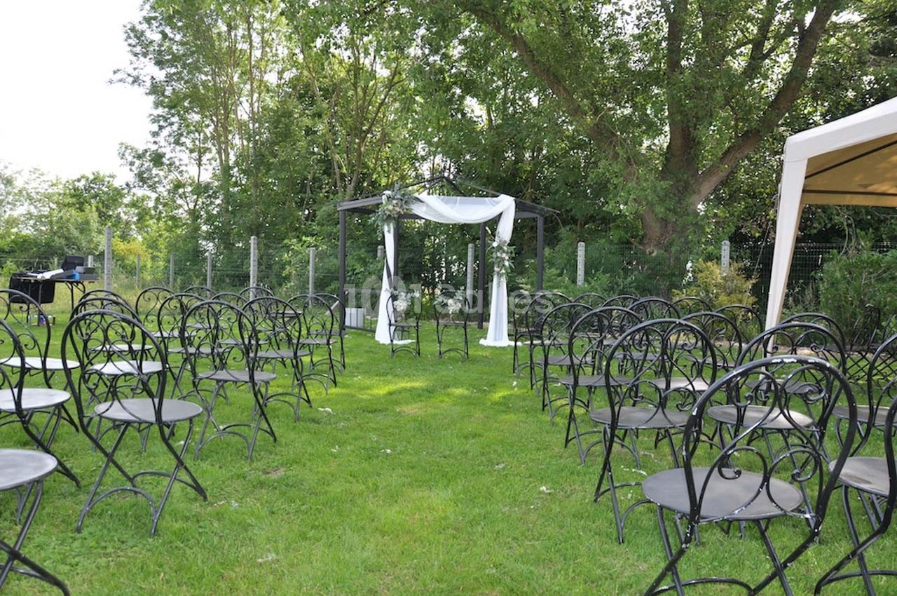 Chaises noires disposées en rangées sur une pelouse, face à une arche décorée de tissu blanc, entourée d'arbres.