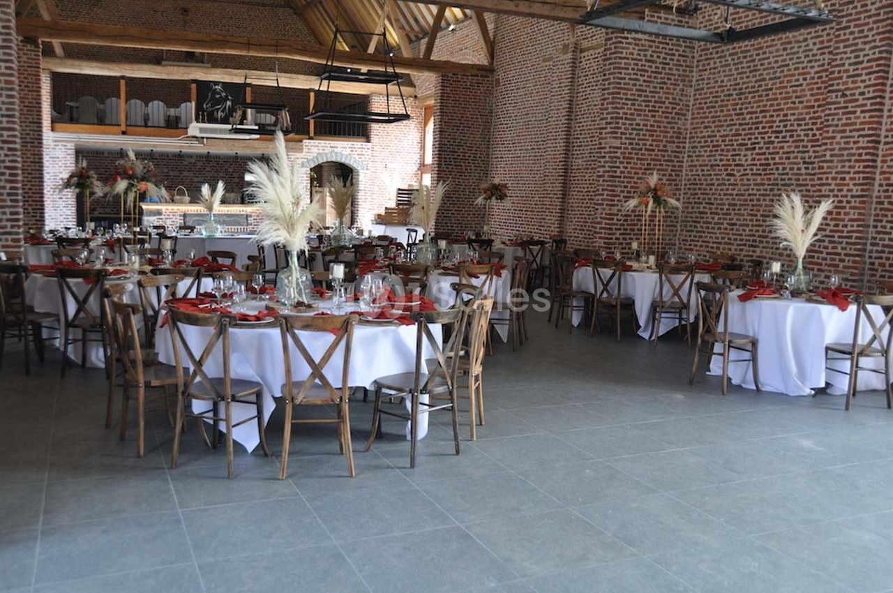 Salle de réception avec tables rondes dressées, nappes blanches, chaises en bois et décorations florales.