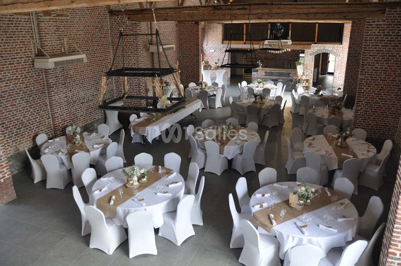 Salle de réception en briques avec tables rondes décorées de nappes blanches et chemins de table en toile de jute.