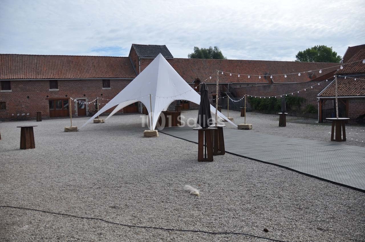 Cour extérieure avec une tente blanche, des tables hautes en bois et des guirlandes lumineuses suspendues.