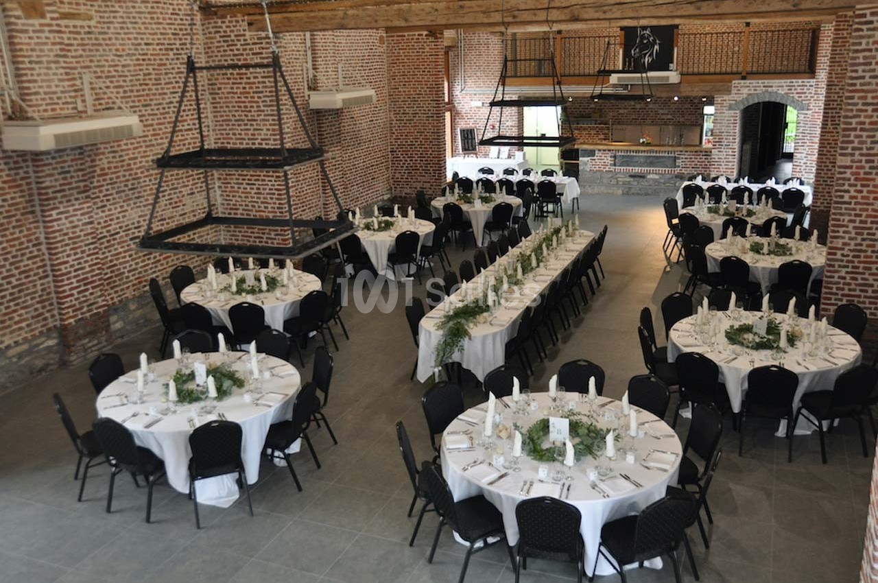 Salle de réception en briques avec tables rondes et rectangulaires dressées pour un événement.