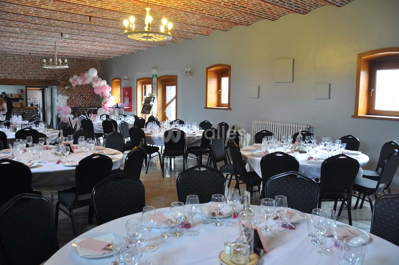 Salle de réception décorée avec des tables rondes dressées, des chaises noires et une arche de ballons roses au fond.