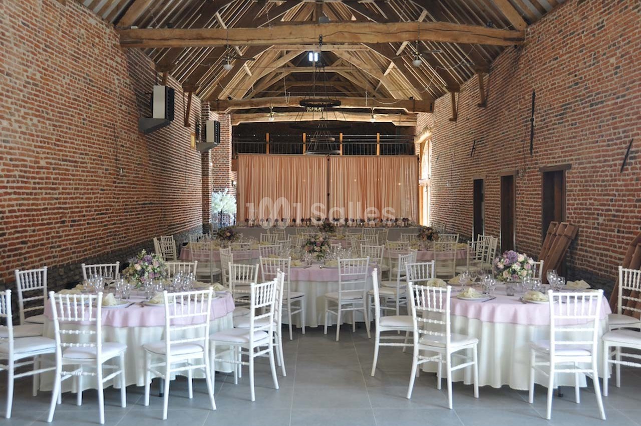 Salle de réception en briques avec poutres apparentes, tables rondes dressées et chaises blanches.