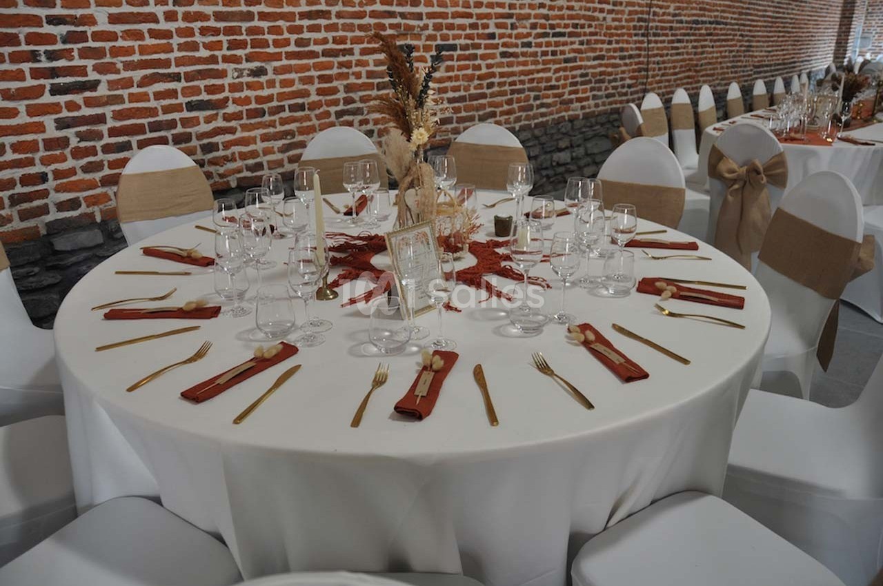 Table ronde décorée pour un événement, avec nappes blanches, couverts dorés, serviettes rouges et centre de table floral.