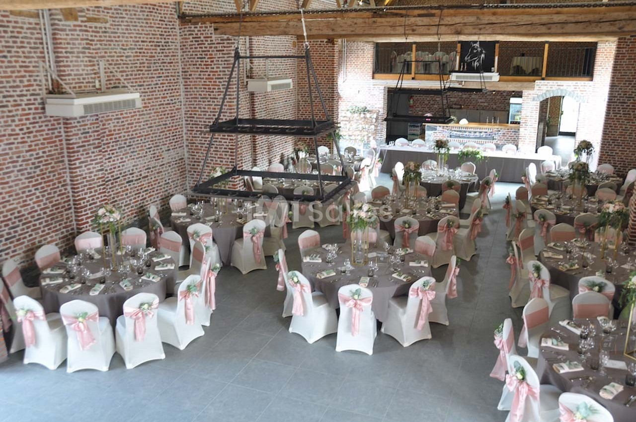 Salle de réception décorée avec des tables rondes, nappes blanches et chaises ornées de rubans roses.