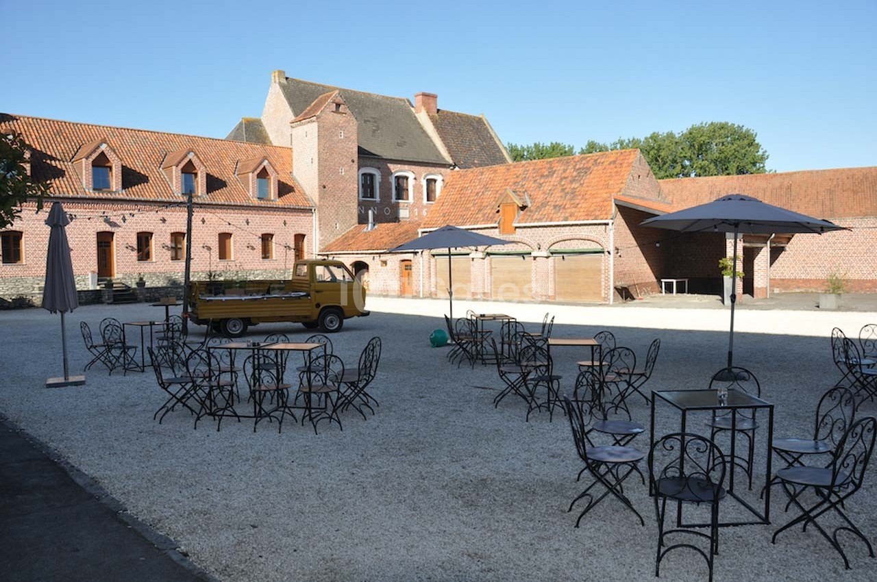 Cour pavée avec des tables et chaises en fer forgé, parasols ouverts, et un bâtiment en briques rouges en arrière-plan.