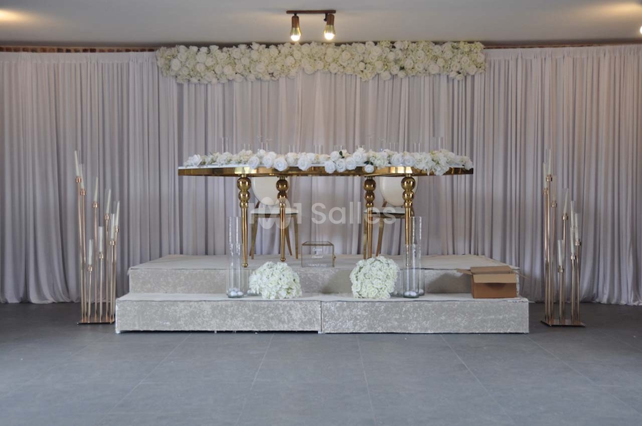 Table décorée avec des fleurs blanches sur une estrade, entourée de rideaux clairs et de chandeliers modernes.