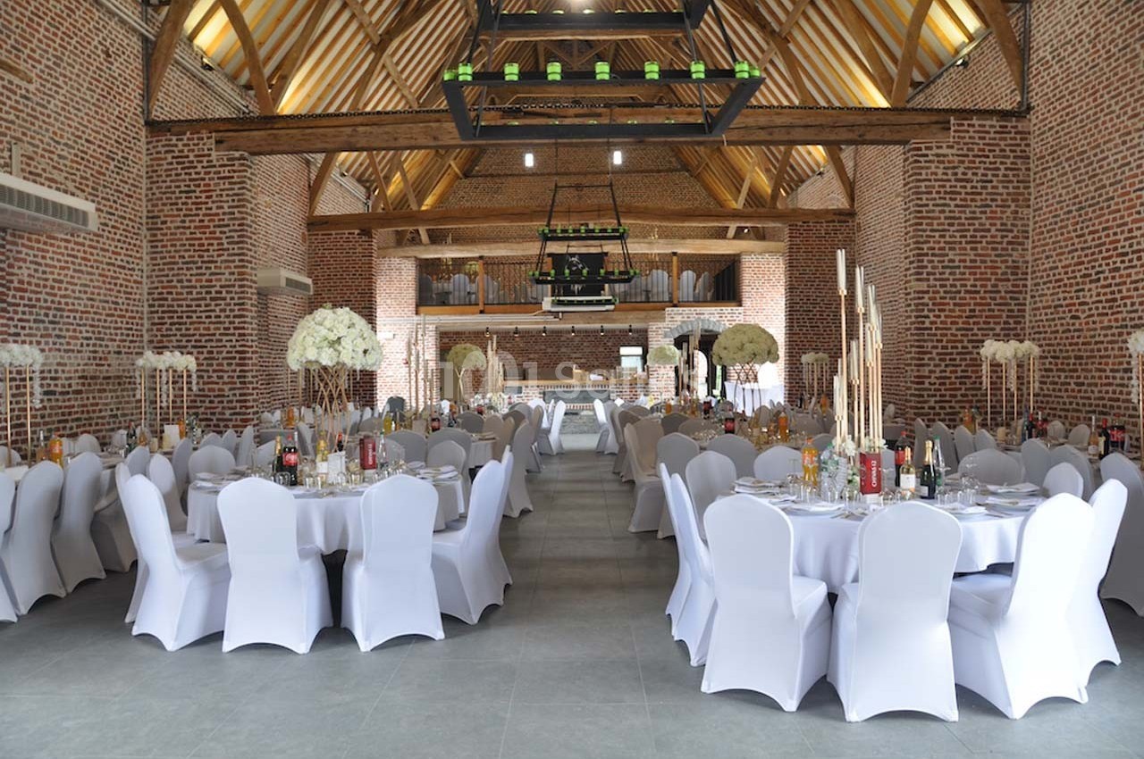 Salle de réception avec tables rondes dressées, chaises blanches et décoration florale, dans un cadre en briques et poutres…