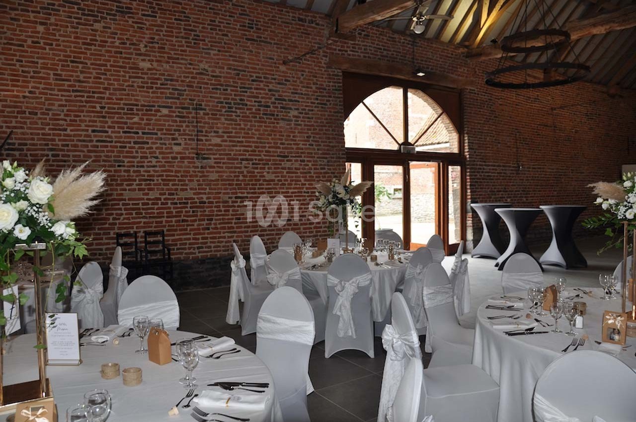 Salle de réception avec murs en briques, tables rondes dressées, chaises blanches et grandes ouvertures vitrées.
