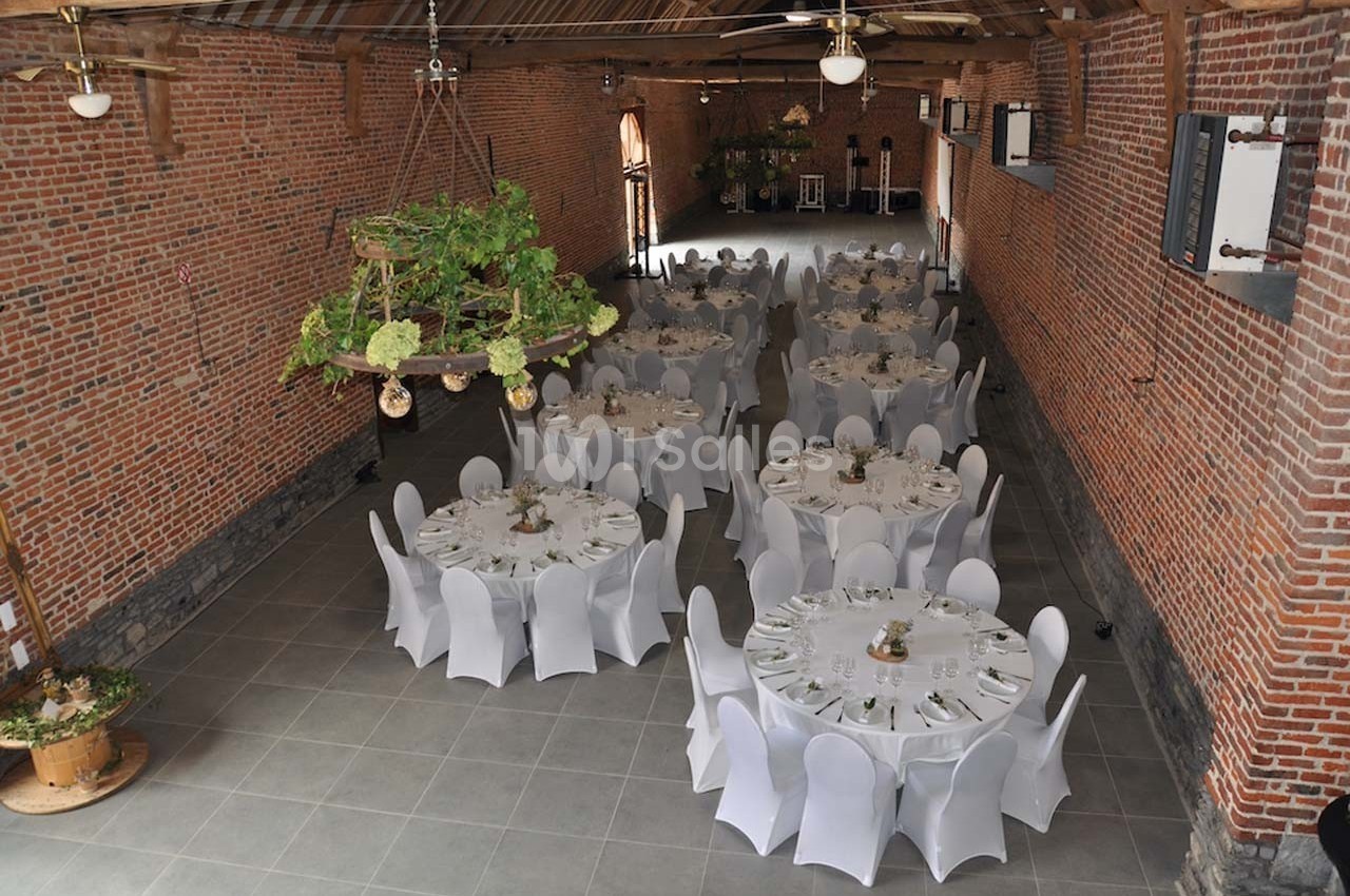Salle de réception en briques avec tables rondes dressées et chaises blanches, décorée de plantes suspendues.