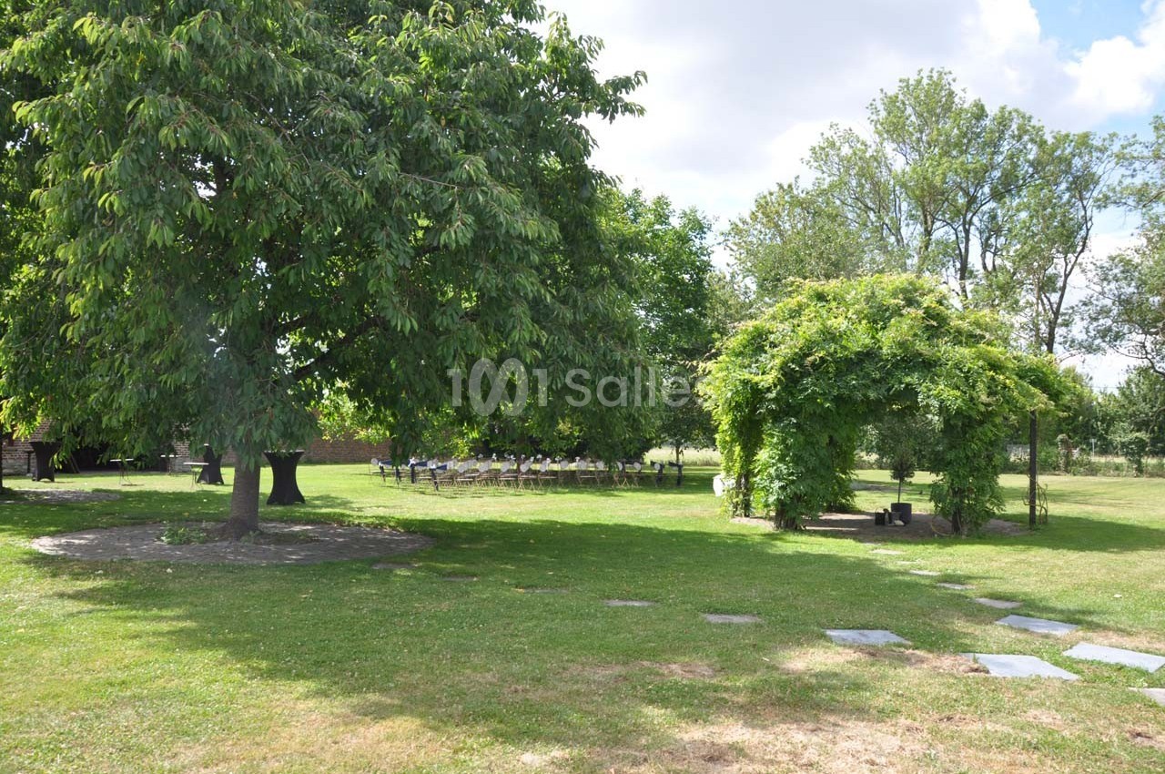 Jardin verdoyant avec un grand arbre, une tonnelle couverte de végétation et des chaises disposées en arrière-plan.