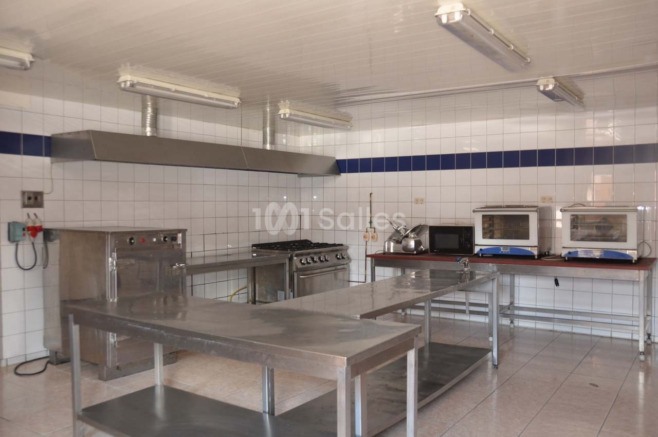 Cuisine professionnelle équipée avec plans de travail en inox, fours, plaques de cuisson et carrelage mural blanc.