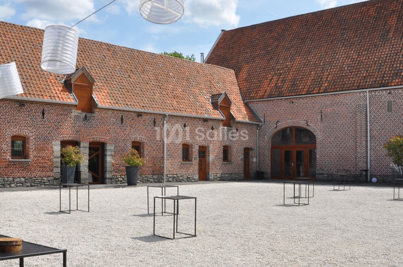Cour pavée avec des bâtiments en briques rouges, des tables métalliques et des lanternes suspendues sous un ciel…