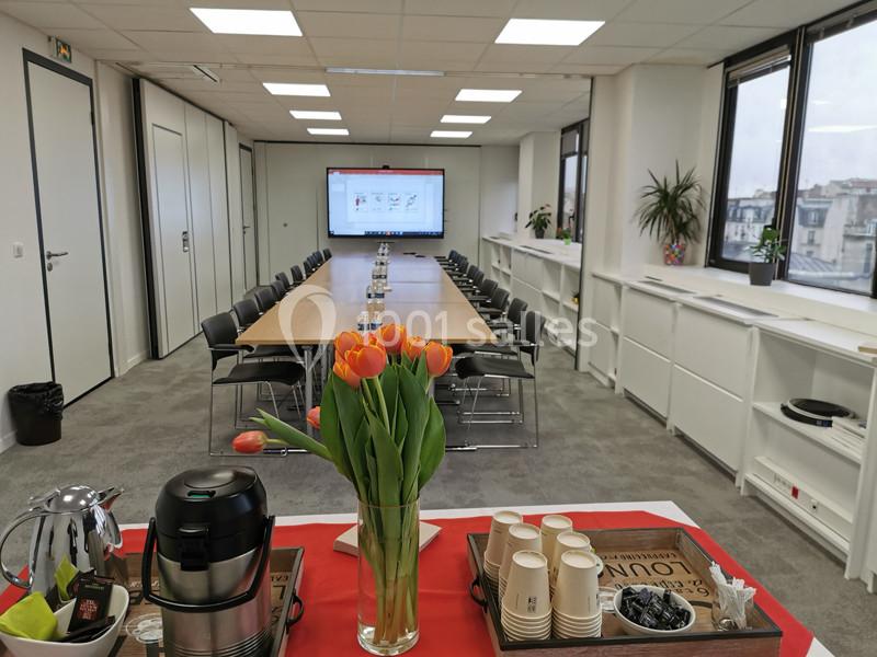 Salle de réunion lumineuse avec une grande table, écran de présentation, et un buffet avec café et fleurs au premier plan.