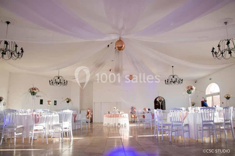 Salle de réception lumineuse décorée de voilages blancs, chaises blanches et tables dressées pour un événement.