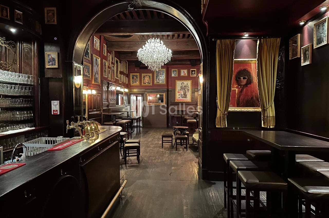 Intérieur d'un bar chaleureux avec comptoir en bois, lustre central, tableaux encadrés et ambiance tamisée.