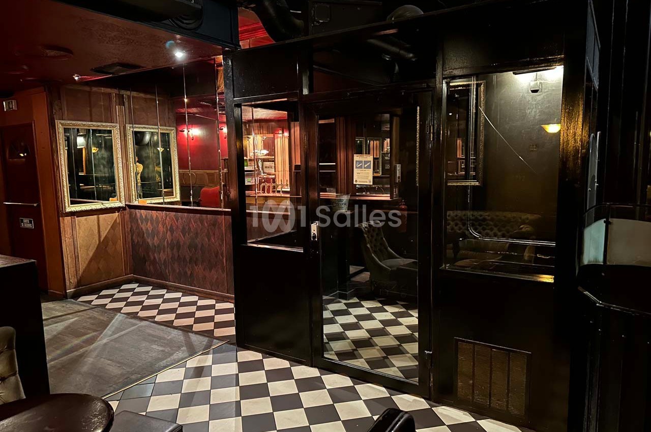 Intérieur d'un bar avec sol en damier, murs sombres, éclairage tamisé et mobilier en bois et cuir.