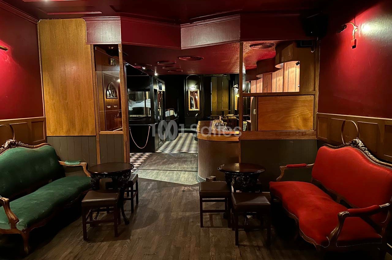 Intérieur d'un salon avec fauteuils rouges et verts, tables basses en bois, éclairage tamisé et sol en parquet.
