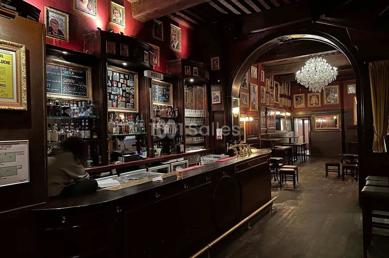 Intérieur d'un bar chaleureux avec comptoir en bois, murs décorés de cadres et lustre suspendu au plafond.