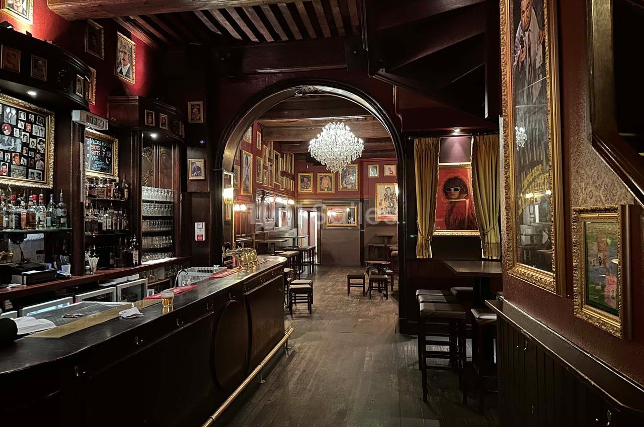 Intérieur d'un bar chaleureux avec un comptoir en bois, des murs décorés de cadres et un lustre central.
