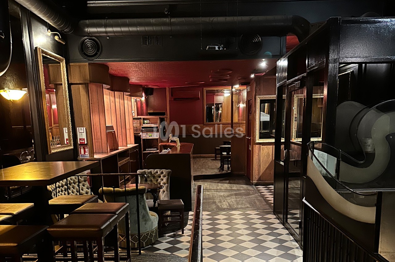 Intérieur d'un bar avec mobilier en bois, fauteuils, sol à carreaux et éclairage tamisé.