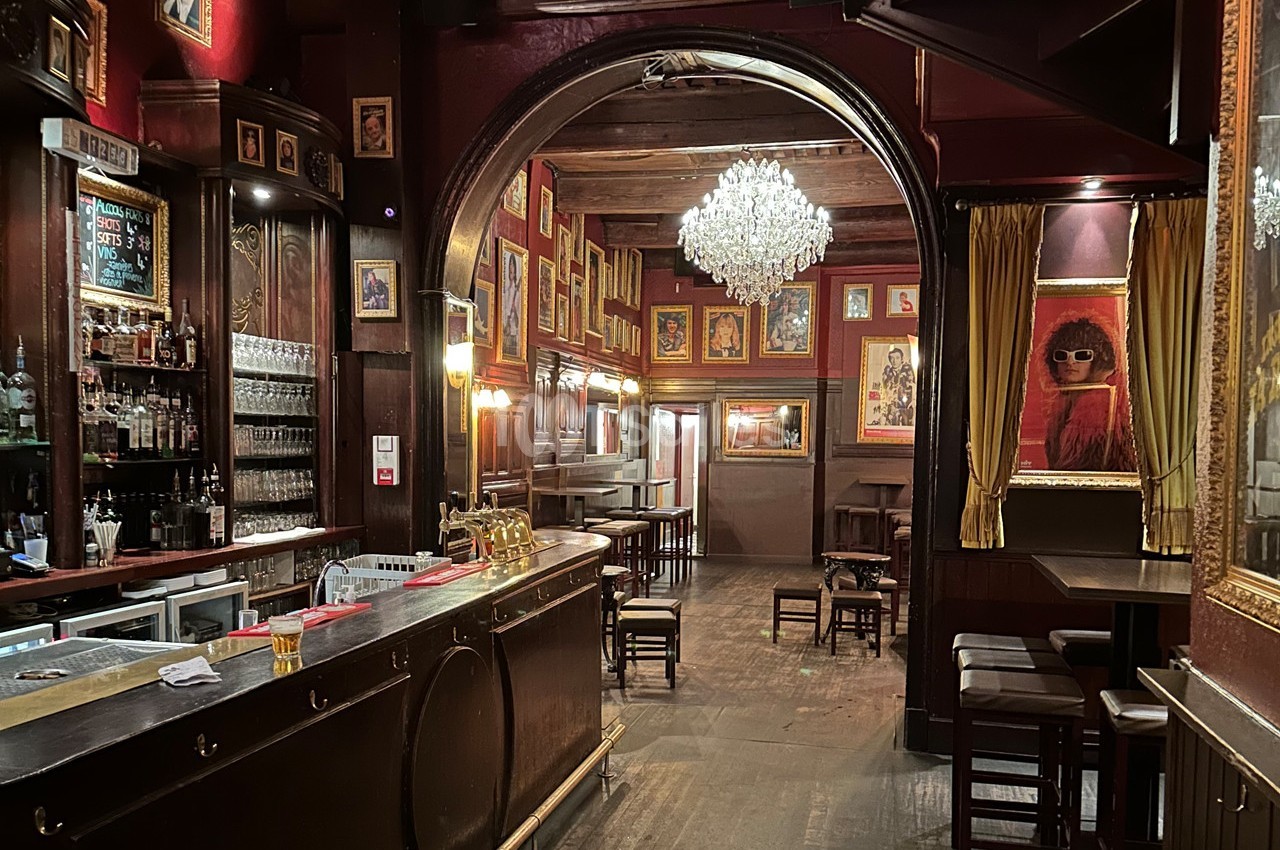 Intérieur d'un bar chaleureux avec comptoir en bois, lustre central, murs rouges décorés de cadres et chaises disposées.