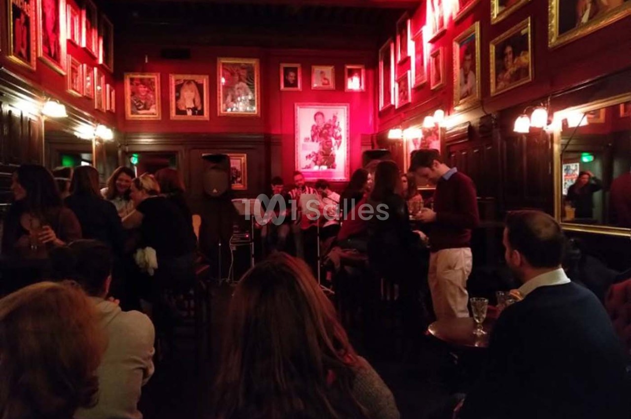 Salle de bar chaleureuse avec éclairage rouge, murs ornés de cadres, et musiciens jouant devant un public assis.