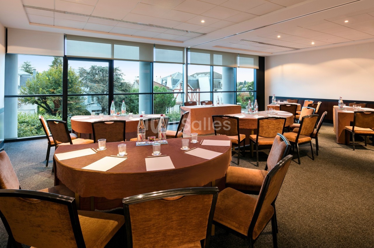Salle de réunion lumineuse avec tables rondes, chaises en bois, grandes baies vitrées et vue sur un jardin.