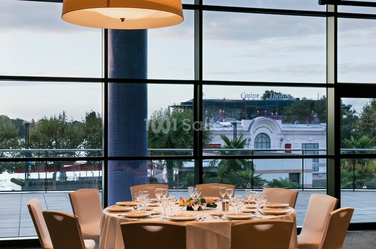 Table dressée avec vaisselle élégante dans une salle lumineuse, vue sur des palmiers et un bâtiment à travers de grandes…