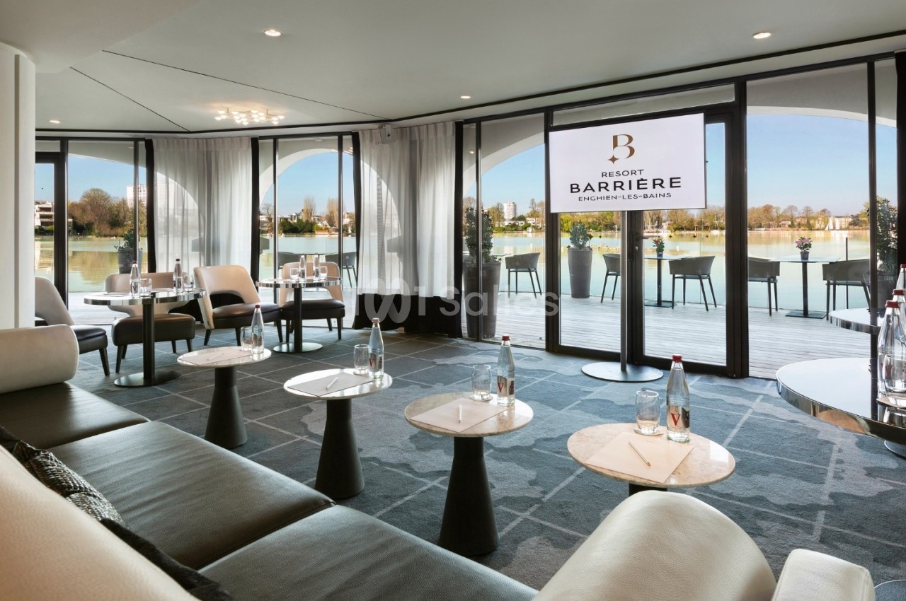 Salon lumineux avec tables basses, fauteuils modernes et vue sur une terrasse donnant sur un plan d'eau.