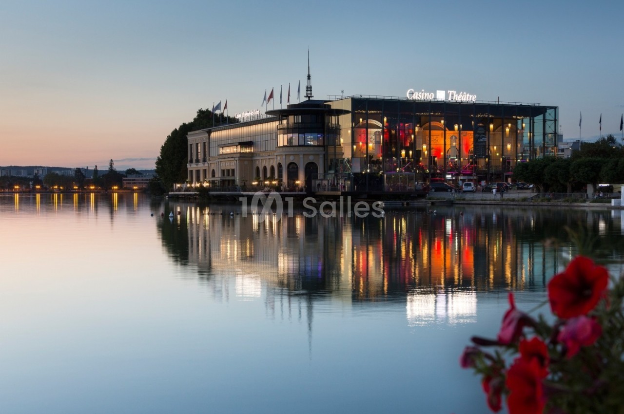 Façade illuminée d'un casino-théâtre au bord d'un lac au crépuscule, avec reflets sur l'eau et fleurs au premier plan.