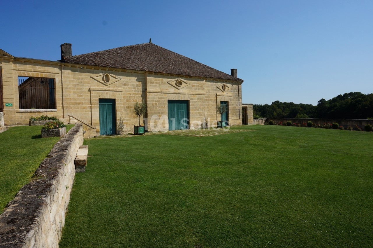 Bâtiment en pierre avec toit en pente, grandes portes vertes et pelouse bien entretenue sous un ciel bleu.
