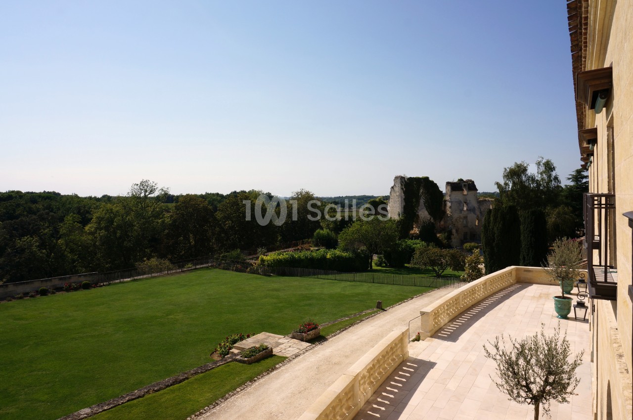 Vue sur un jardin verdoyant avec pelouse, arbres et ruines anciennes, depuis une terrasse en pierre par temps clair.