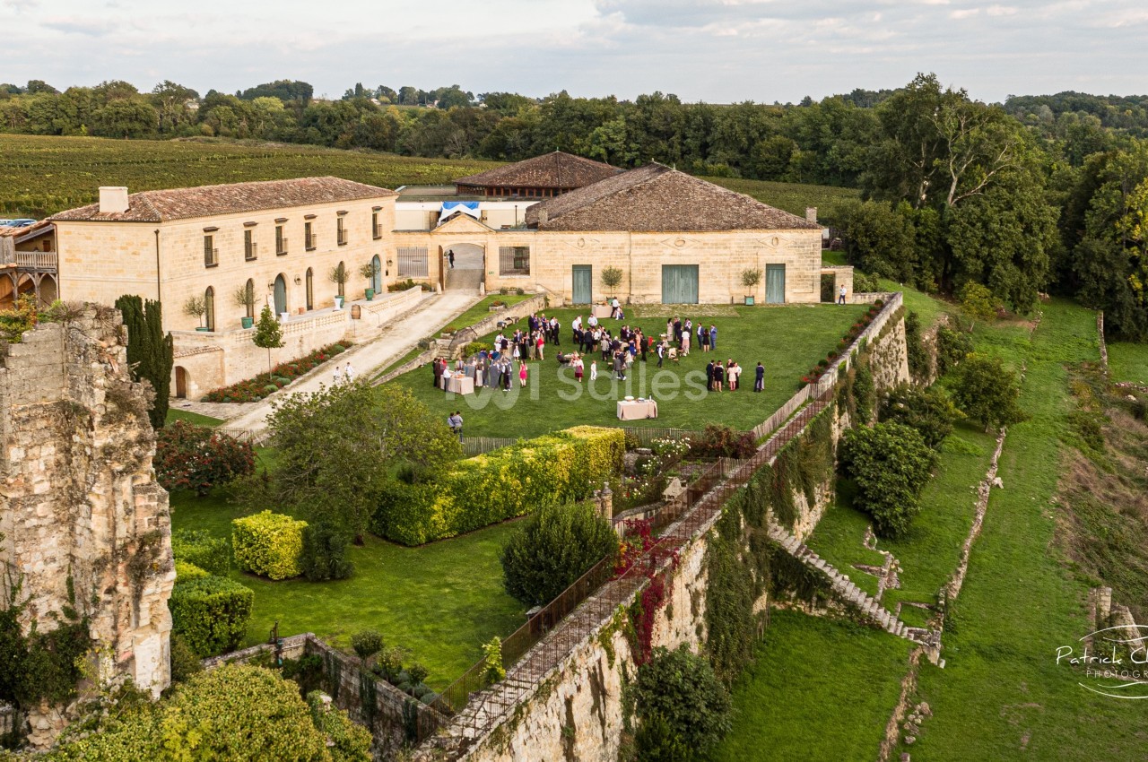 Vue aérienne d'un domaine avec un bâtiment en pierre, un jardin verdoyant et un rassemblement de personnes en extérieur.