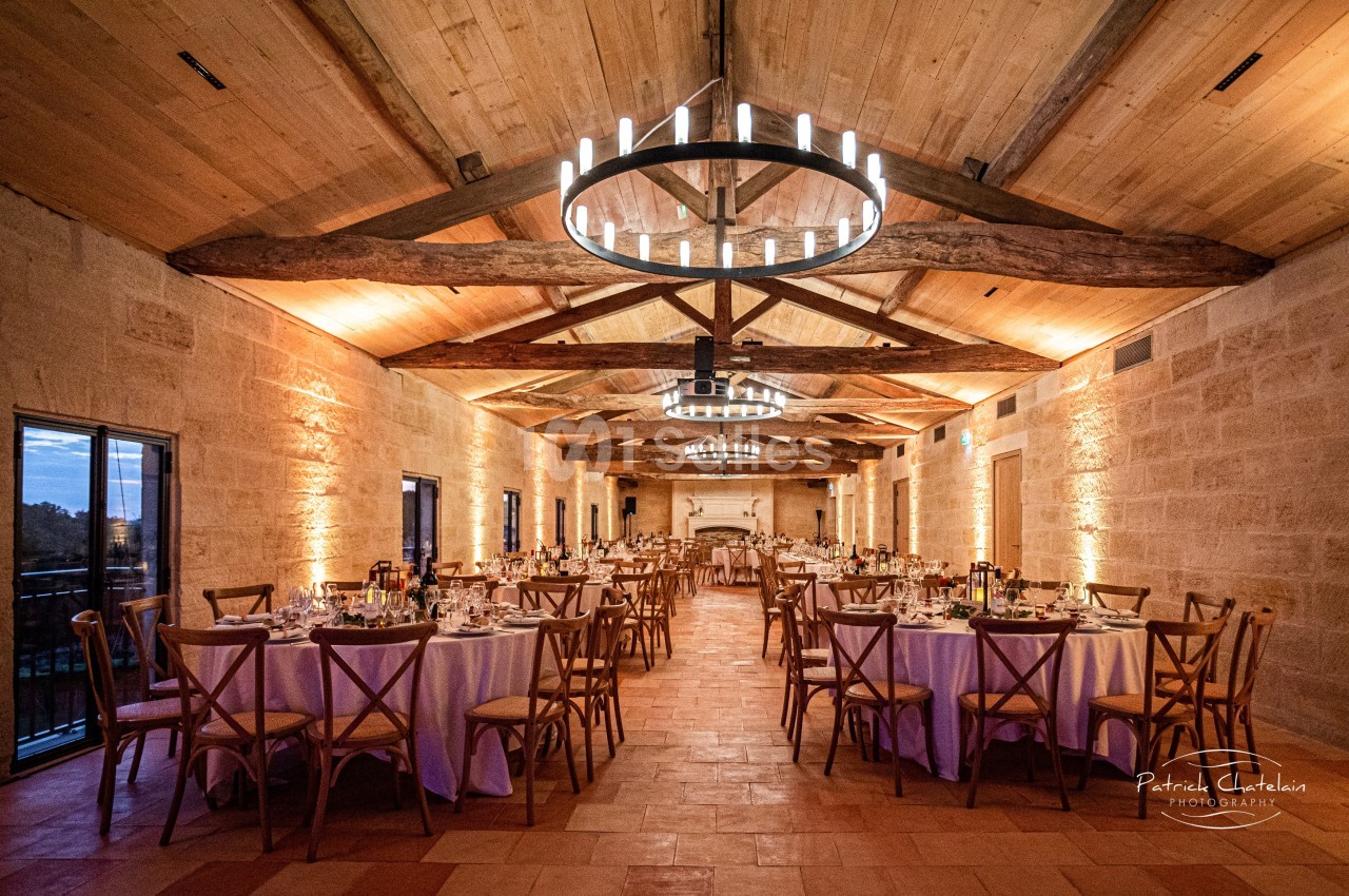 Salle de réception en pierre avec tables dressées, chaises en bois et lustres circulaires suspendus au plafond en poutres…