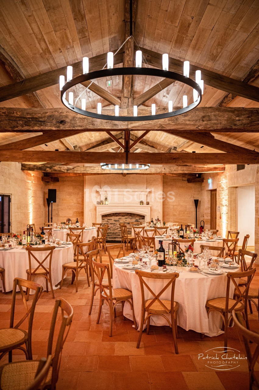 Salle de réception chaleureuse avec tables rondes dressées, chaises en bois et un lustre circulaire suspendu.