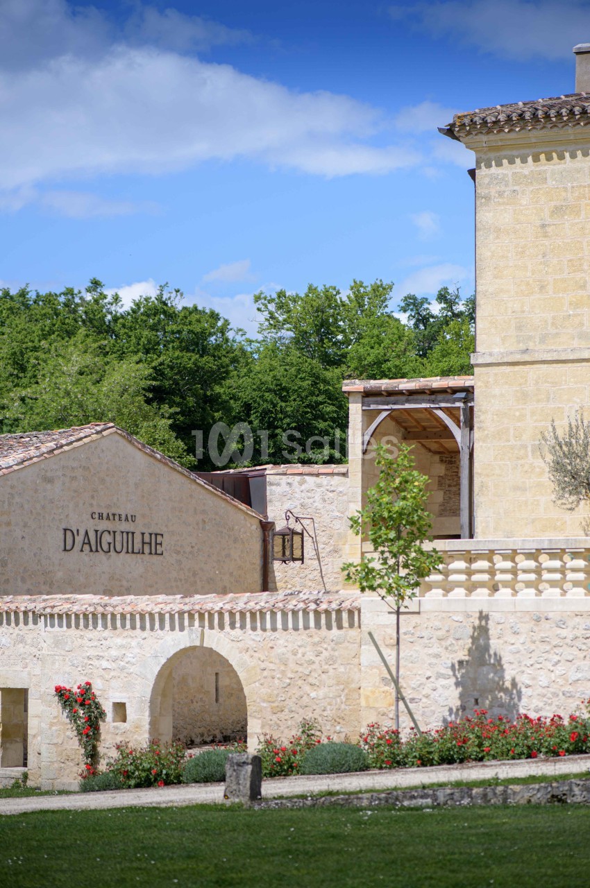 Bâtiments en pierre d'un domaine viticole avec jardin fleuri, sous un ciel bleu partiellement nuageux.