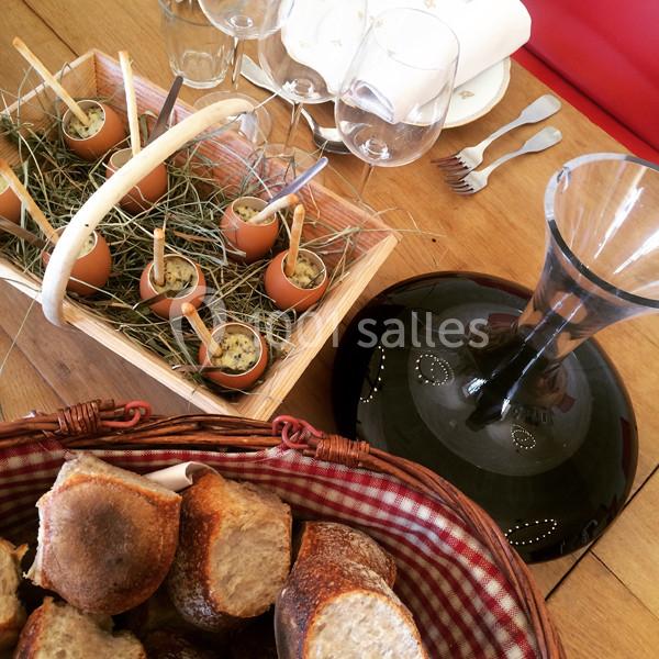 Des œufs servis dans des coquilles sur un lit de foin, avec une carafe de vin et un panier de pain sur une table en bois.
