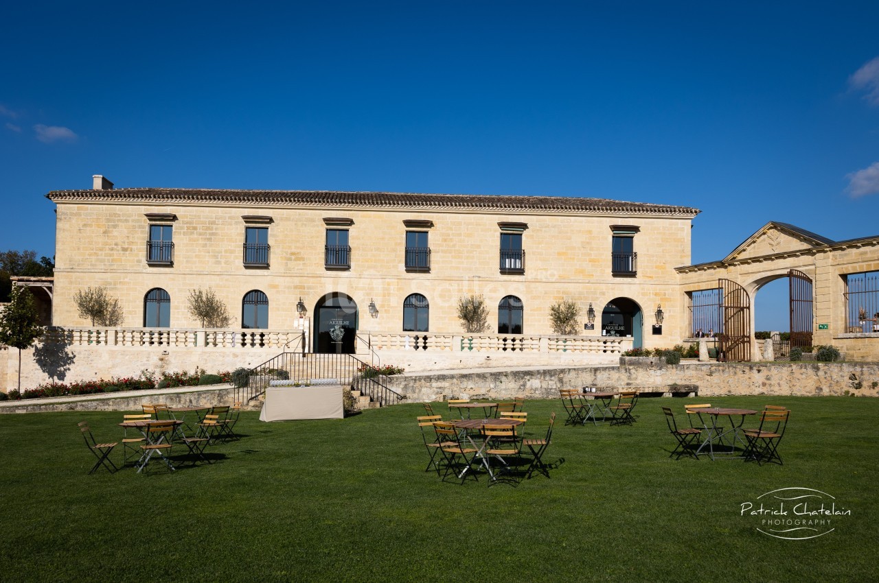 Façade d'un bâtiment en pierre avec des fenêtres noires, entouré de pelouses et de tables en plein air.