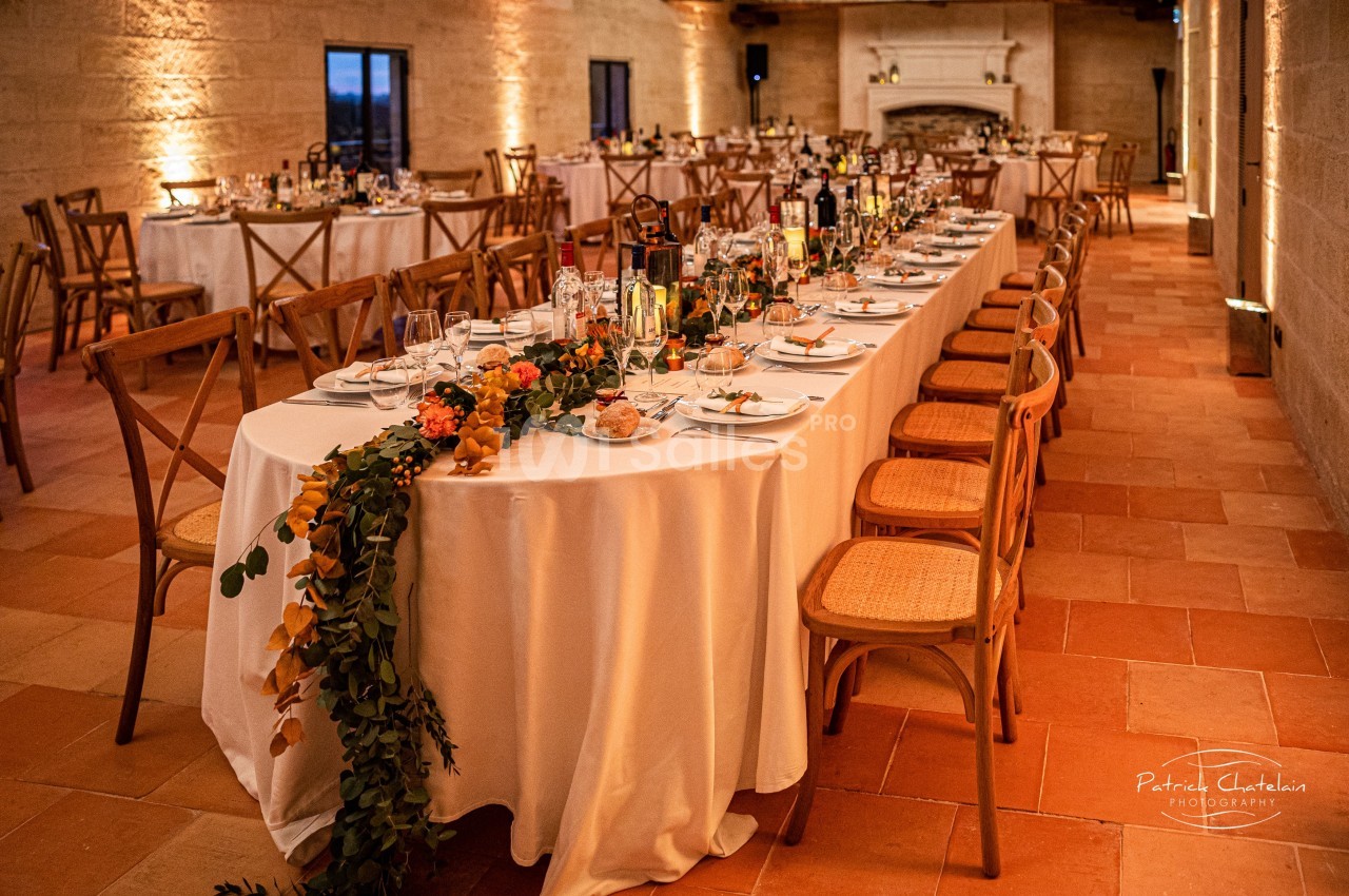 Grande table de réception décorée avec des fleurs et de la vaisselle, dans une salle aux murs en pierre et éclairage…
