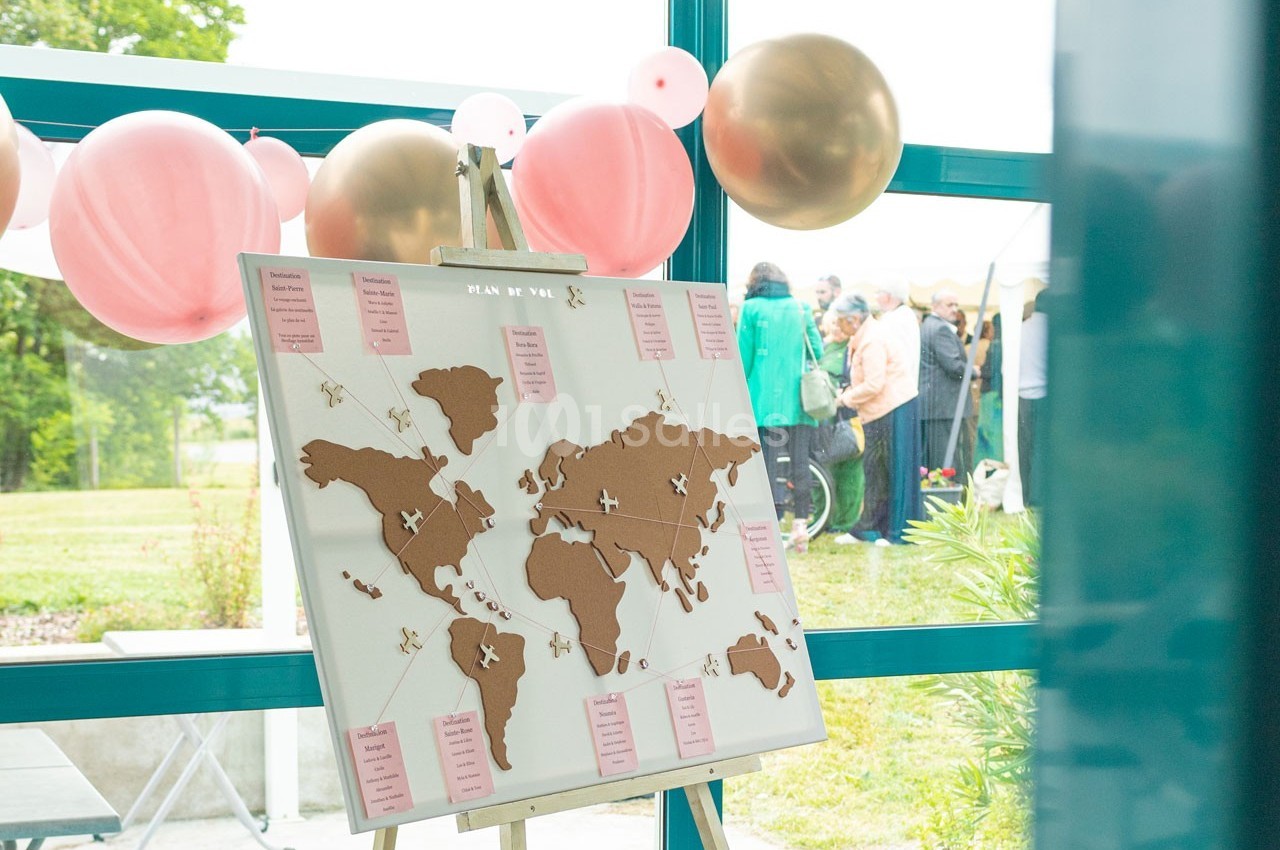 Plan de table en forme de carte du monde avec des marqueurs, entouré de ballons roses et dorés, dans une salle lumineuse.