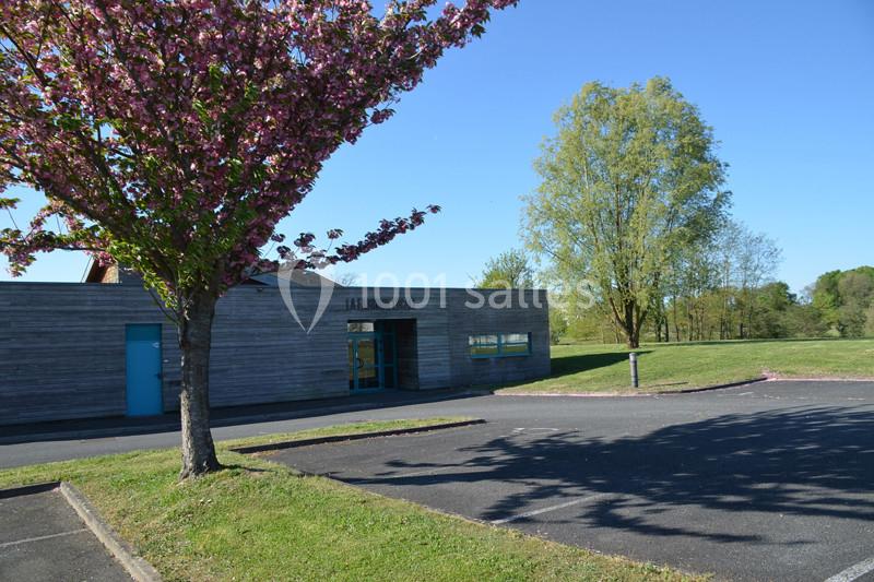 Façade d'un bâtiment en bois entouré d'arbres, avec un parking et une pelouse sous un ciel dégagé.