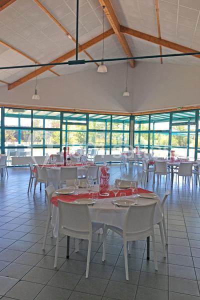 Salle lumineuse avec tables rondes dressées, nappes blanches et décorations rouges, grandes baies vitrées en arrière-plan.