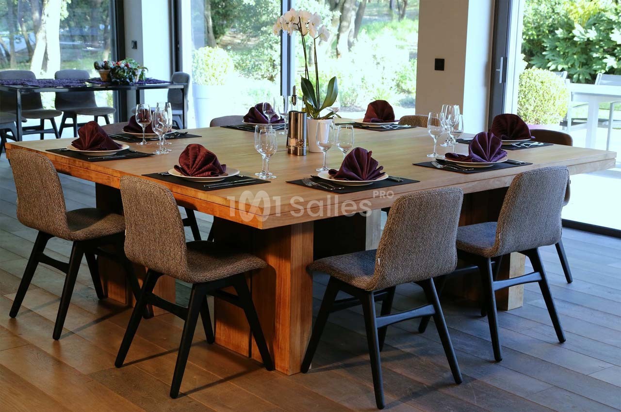 Table en bois dressée avec huit chaises, serviettes pliées violettes, verres et couverts dans une salle lumineuse.